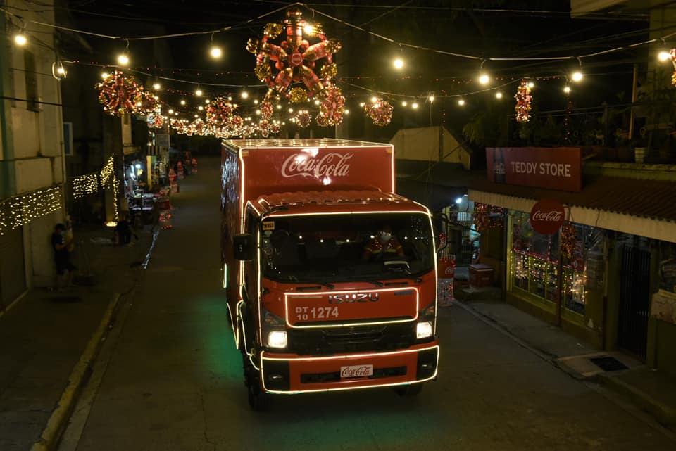 A red Coca-Cola Santa truck makes its way through a street illuminated by Coca-Cola parol lanterns.